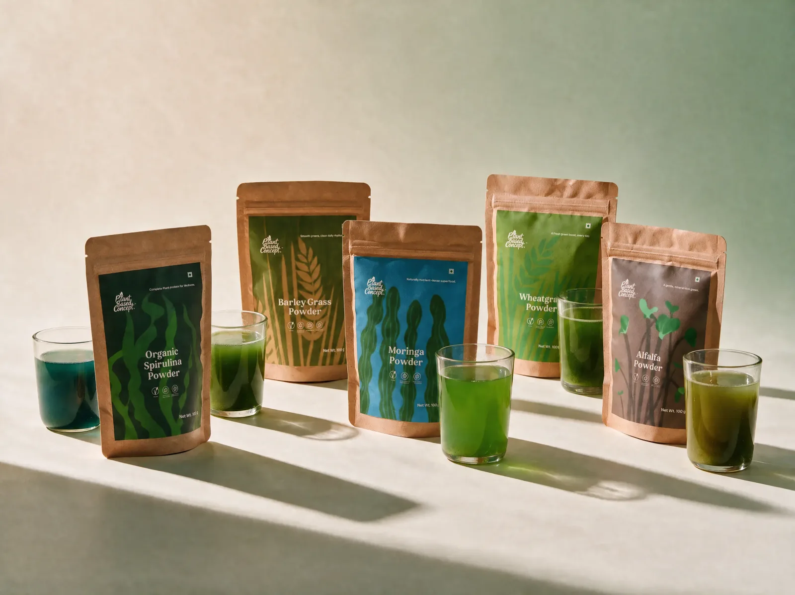 The five Plant Based Concept pouches — spirulina, barley grass, moringa, wheatgrass and alfalfa — each beside a glass of its freshly mixed green drink, on a soft studio surface